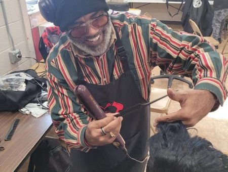 A barber with glasses and headphones is smiling while cutting a student's hair in a school salon.